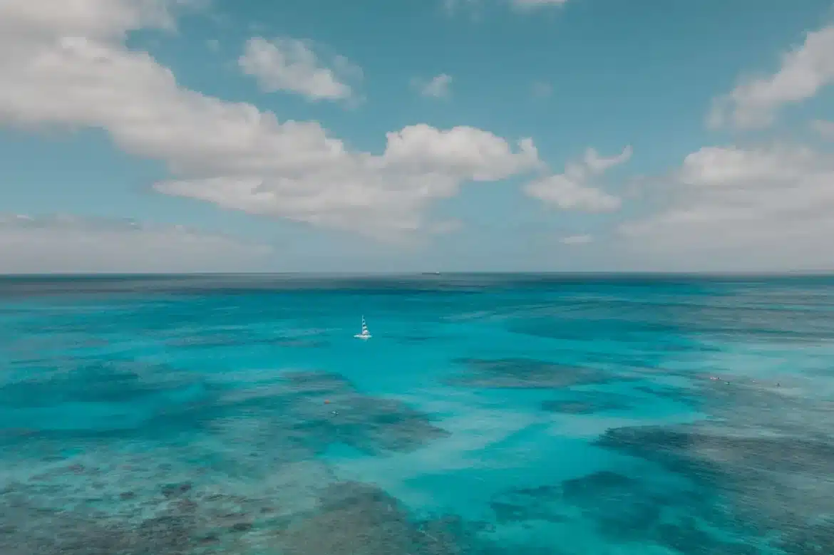 Plage de sable blanc aux eaux turquoise avec un voilier à l'horizon sous le soleil des Caraïbes